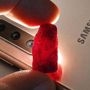 May include: A rough-cut, deep red gemstone is held between two fingers, set against a gold-colored Samsung phone. The gemstone's texture is visible, and the light catches its facets, highlighting its color and shape.