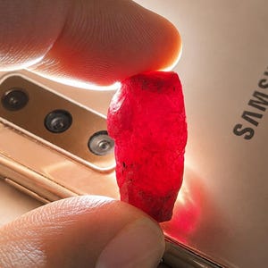 May include: A vibrant red gemstone held between fingers, illuminated against a gold-colored smartphone. The rough-cut ruby displays a deep, saturated color, showcasing its natural texture and form. The phone's camera lenses are visible.
