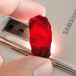 May include: A rough, deep red gemstone is held between fingers, showcasing its natural, irregular shape. The stone's color is a vibrant, saturated red, with a slightly translucent quality. The background is a light-colored phone with the brand name "SAMSUNG" visible.