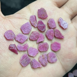 May include: A collection of raw, uncut ruby gemstones in varying shapes and sizes. The rubies display a range of pink to deep red hues, with a rough, textured surface. The gemstones are scattered across a neutral background.