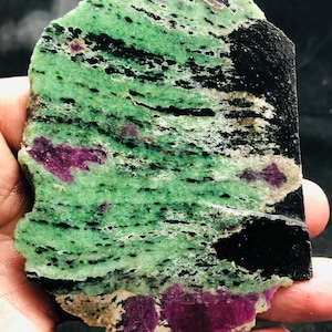 Puede incluir: Una piedra rugosa, verde y negra con inclusiones moradas. La piedra tiene un aspecto estratificado y estriado.