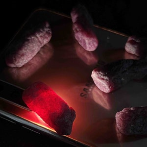 May include: A collection of rough, uncut gemstones in varying sizes and shapes, with a deep red hue. The gems are illuminated, creating a glowing effect. The background is a reflective surface, possibly a phone.