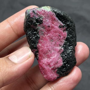 May include: A raw, unpolished mineral specimen featuring a vibrant, textured magenta-pink centre surrounded by a rough, dark black outer crust. The stone has an irregular, organic shape with visible crystalline formations and a glossy surface finish.