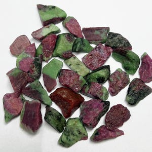 May include: A collection of raw ruby zoisite stones. The stones display a mix of colours, including deep red, green, and a mottled appearance. The rough-cut gemstones are scattered on a white surface.