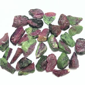 May include: A collection of raw ruby and zoisite stones. The stones display a mix of colors, including deep red, green, and black. The rough, natural texture of the stones is visible, with varying shapes and sizes scattered across a white surface.