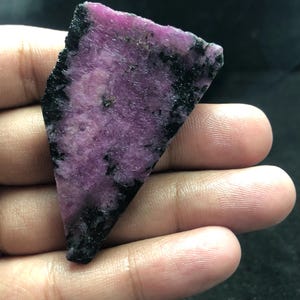 Puede incluir: Una rebanada de piedra preciosa en bruto de forma triangular que presenta un centro magenta rosado vibrante con inclusiones minerales negras profundas a lo largo de los bordes y en toda la superficie, con una textura rugosa y sin pulir.