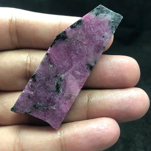 Puede incluir: Una gema rectangular en bruto con un vibrante tono rosa, acentuada por inclusiones negras. La piedra tiene una textura natural rugosa y se sostiene en una mano, mostrando su apariencia única y sin pulir. Un ejemplo llamativo de belleza mineral natural.