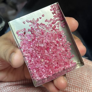 May include: A small, rectangular metal tray filled with numerous small, irregular-shaped pink gemstones. The gemstones vary in shade from light to deep pink. The tray is held by a person, with the background slightly blurred.