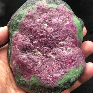 Puede incluir: Una gema de rubí en zoisita en bruto. La piedra presenta un vibrante centro magenta, rodeado por un borde verde y negro. La piedra natural se sostiene en una mano, mostrando su tamaño y textura.
