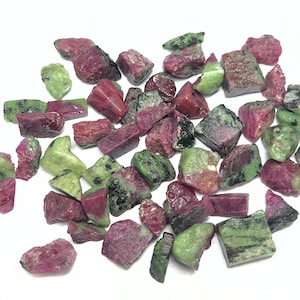 May include: A selection of raw ruby and zoisite gemstones. The stones exhibit a combination of deep red and green colours, with a natural, unpolished texture. The gemstones are spread across a white surface, highlighting their diverse shapes and sizes.
