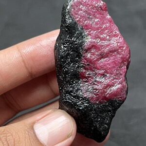 Peut inclure: Un spécimen minéral brut et non poli présentant un contraste saisissant entre une section noire profonde et brillante et une partie cristalline texturée d'un rose magenta vibrant. La pierre a une forme irrégulière et dentelée.