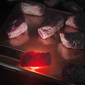 May include: A collection of pink and red gemstones, some of which are illuminated by a light source, on a metallic surface.