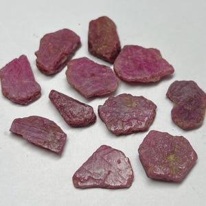 May include: A collection of rough-cut ruby gemstones in varying shapes and sizes. The stones display a deep pinkish-red hue, with visible textures and natural imperfections. The gemstones are scattered on a white surface.