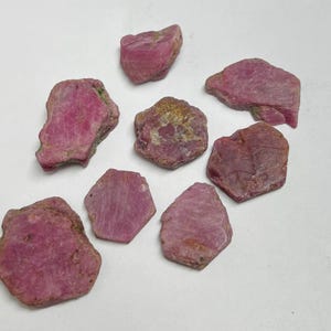 May include: A collection of rough, uncut ruby gemstones. The stones vary in size and shape, displaying a deep pink to reddish-pink hue. Some stones have a slightly rough, textured surface, while others appear smoother.