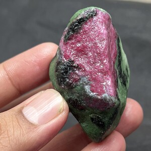 May include: A rough, green and pink gemstone with black inclusions. The gemstone is irregularly shaped and has a smooth, polished surface.