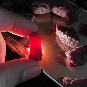 Puede incluir: Primer plano de una colección de piedras preciosas rojas, algunas de las cuales están siendo sostenidas por una mano. Las piedras preciosas son brillantes y tienen una textura rugosa y natural.