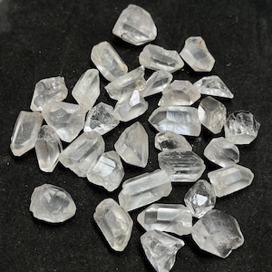 Può includere: Una collezione di piccoli cristalli di quarzo grezzi, trasparenti, sotto forma di punte e frammenti. Ogni pezzo presenta sfaccettature geometriche naturali e irregolari con vari gradi di trasparenza e inclusioni interne sparse nel quadro.