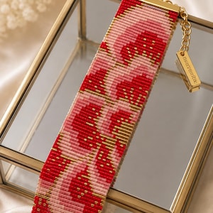 May include: A wide beaded bracelet featuring a repeating pattern of red, light pink, and metallic gold seed beads. The bracelet has gold-toned metal end bars and a gold chain extension with a rectangular metal tag engraved with the text marpunitik.