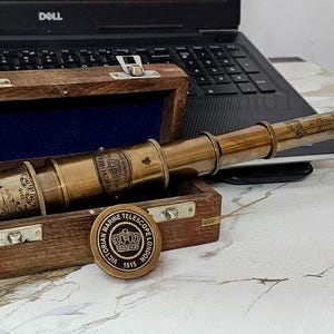 May include: A vintage brass telescope with a wooden case. The telescope is engraved with text and rests inside an open wooden box lined with blue velvet. A brass medallion with a crown is next to the box. A laptop is in the background.