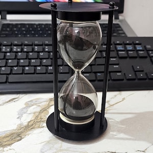 May include: A black hourglass with a clear glass center filled with black sand. The hourglass is encased in a black metal frame with a circular base and top. The timekeeping device is on a white and gray marble surface.