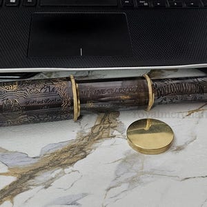 May include: A vintage-style brass telescope with intricate engraved patterns and gold accents. The telescope is dark brown with gold rings and end caps. A separate gold-colored base is also present. The telescope is resting on a marble surface.