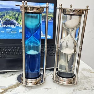 May include: Two hourglasses with silver-colored metal frames. One hourglass contains blue liquid and white sand, the other has white sand. The hourglasses are on a white surface, with a laptop in the background.