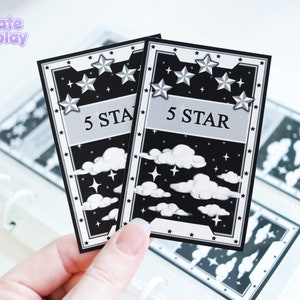 Pode incluir: Dois cartões preto e branco com uma borda branca e um fundo preto. Os cartões têm uma faixa branca com o texto "5 STAR" e cinco estrelas prateadas. O fundo dos cartões tem nuvens brancas e estrelas prateadas.