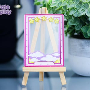 May include: A pink and purple card with a white frame and yellow stars. The card is on a wooden easel and has a cloud design.