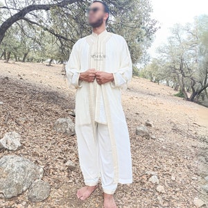 May include: A white robe with gold embroidery and a white undershirt and pants. The robe has a collar and is worn by a person standing in a forest setting.