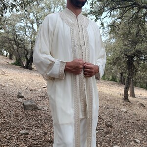 May include: A white long-sleeved robe with intricate embroidery and a button closure. The robe is worn by a person standing in a forest setting.
