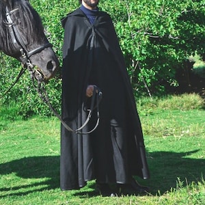 May include: A person wearing a black hooded cloak stands next to a black horse with a leather bridle. The person is holding a black leather strap.