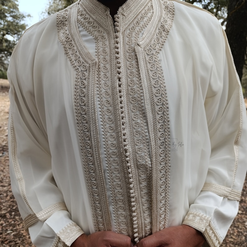 May include: A white long-sleeved shirt with intricate gold embroidery and a button-up closure. The shirt has a high collar and a traditional design.