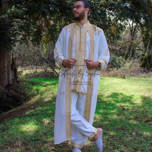 May include: A white traditional garment with gold embroidery, including a long tunic and matching pants. The outfit features intricate gold detailing along the edges and a zipper closure. The text "Softouch By Sophie" is visible on the garment.