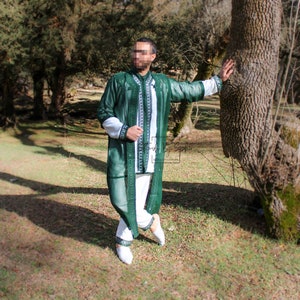 May include: A green, long-sleeved garment with white accents and intricate embroidery. The garment is worn over white pants and a white shirt. The outfit is completed with white shoes. The image is taken outdoors with trees in the background.