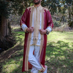 May include: A burgundy and gold embroidered robe with a white tunic and pants. The outfit has gold trim and embroidery on the sleeves and collar. The ensemble is worn with light blue slippers. The text "Stonish By Soph" is visible on the tunic.