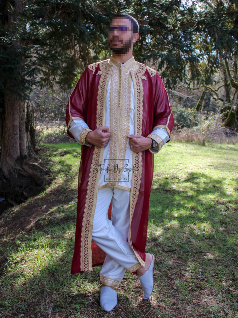 May include: A burgundy and gold embroidered robe with a white tunic and pants. The outfit has gold trim and embroidery on the sleeves and collar. The ensemble is worn with light blue slippers. The text "Stonish By Soph" is visible on the tunic.