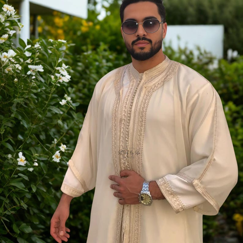 May include: Cream-colored, long-sleeved garment with intricate gold embroidery along the front and sleeves. The garment has a mandarin collar and a relaxed fit. The person is wearing sunglasses and a silver watch.