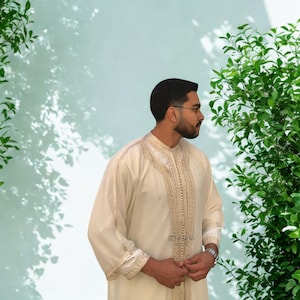 May include: Cream-colored traditional garment with intricate gold embroidery along the collar, front, and cuffs. The long-sleeved robe-like attire features a high neckline and a subtle sheen. The garment is worn by a person with dark hair and a beard.