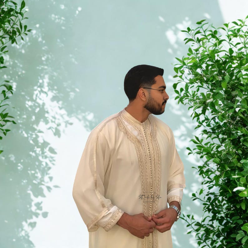 May include: Cream-colored traditional garment with intricate gold embroidery along the collar, front, and cuffs. The long-sleeved robe-like attire features a high neckline and a subtle sheen. The garment is worn by a person with dark hair and a beard.