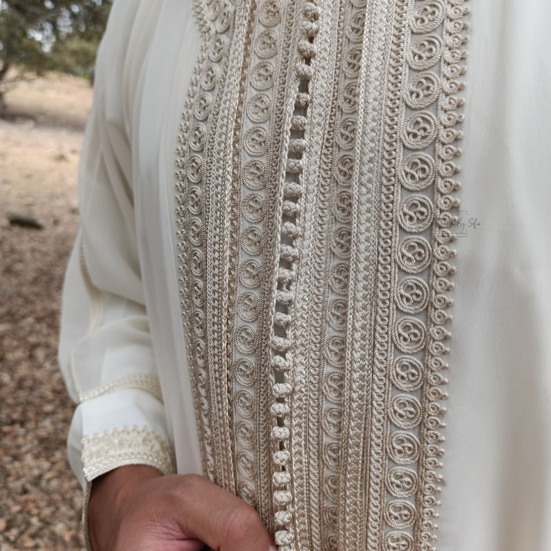 May include: A white long-sleeved garment with intricate gold embroidery along the front. The embroidery features a repeating pattern of circles and loops.