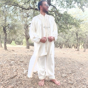 May include: A white traditional robe with gold embroidery and a matching white pants set. The robe has a long, flowing design and is worn by a person standing in a forest setting.