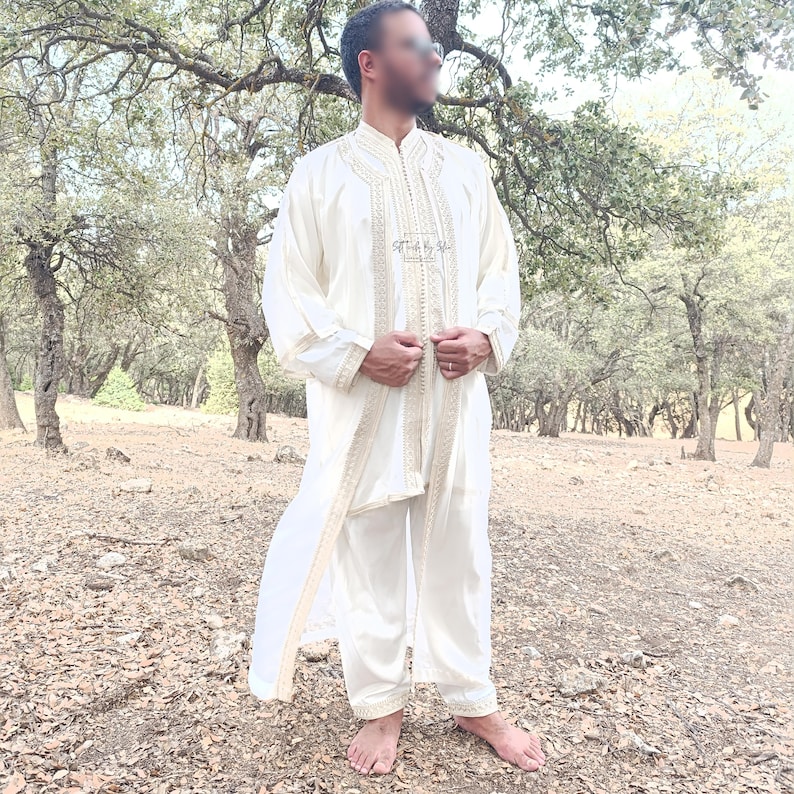 May include: A white traditional robe with gold embroidery and a matching white pants set. The robe has a long, flowing design and is worn by a person standing in a forest setting.