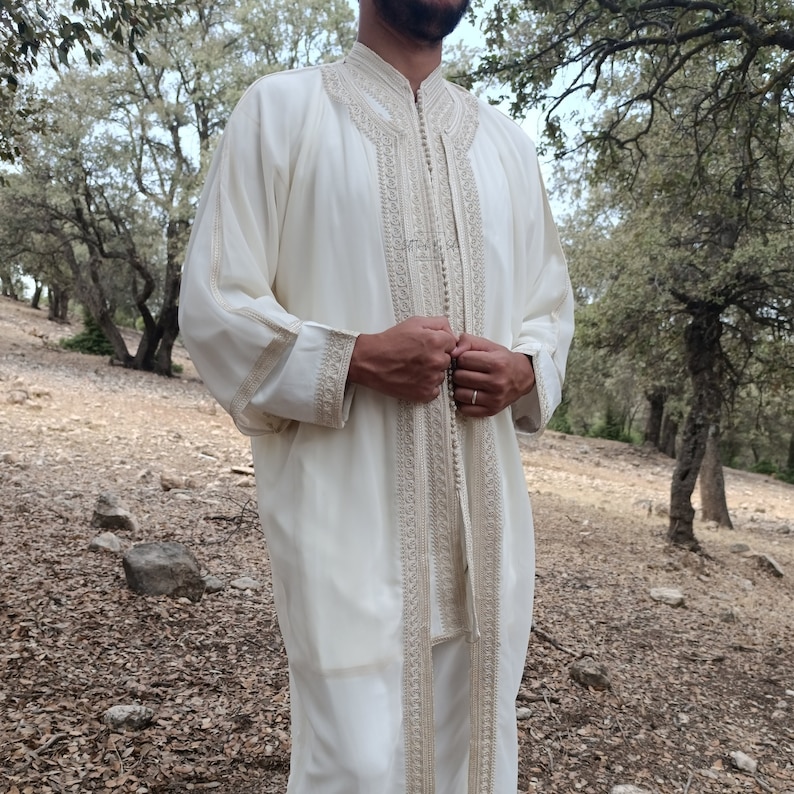 May include: A white long-sleeved robe with intricate embroidery and a button closure. The robe is worn by a person standing in a forest setting.
