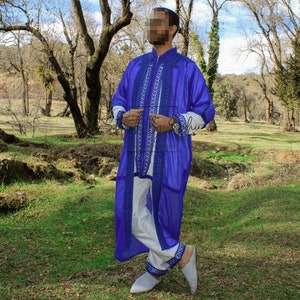 May include: A vibrant blue robe with intricate white and blue embroidery, paired with white pants and matching embroidered trim. The outfit includes white slippers. The ensemble is set against a natural outdoor backdrop.