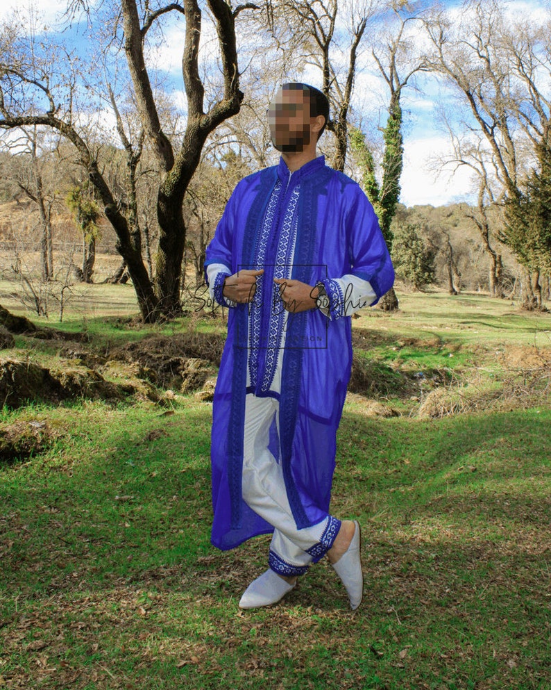 May include: A vibrant blue robe with intricate white and blue embroidery, paired with white pants and matching embroidered trim. The outfit includes white slippers. The ensemble is set against a natural outdoor backdrop.