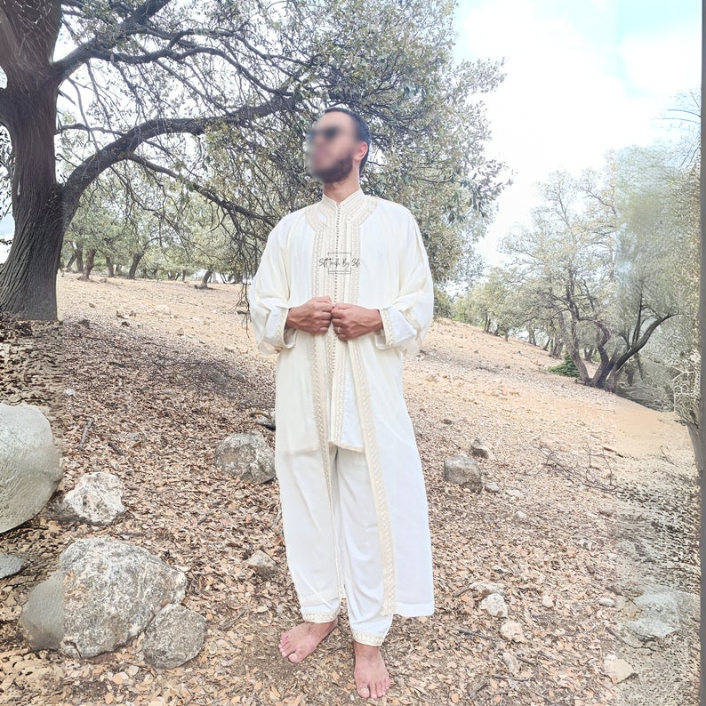 May include: A white robe with gold embroidery and a white undershirt and pants. The robe has a collar and is worn by a person standing in a forest setting.