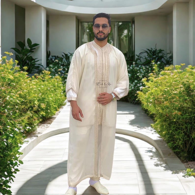 May include: Cream-colored, long-sleeved garment with intricate gold embroidery. The outfit includes matching pants and shoes. The garment has a button-down front and is worn by a person wearing sunglasses and a watch. The text "S&T by Sh-" is visible on the garment.