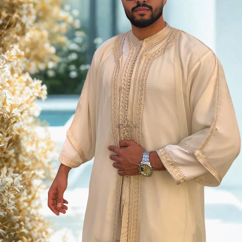 May include: Cream-colored traditional garment with intricate gold embroidery along the front and sleeves. The long-sleeved robe features a mandarin collar and a flowing design. The garment is worn by a person with a beard and glasses.