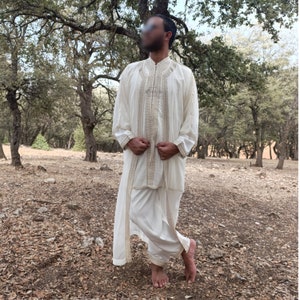 May include: A white long-sleeved robe with gold embroidery and a matching white pant. The robe is open in the front and the person is wearing it with the sleeves rolled up.