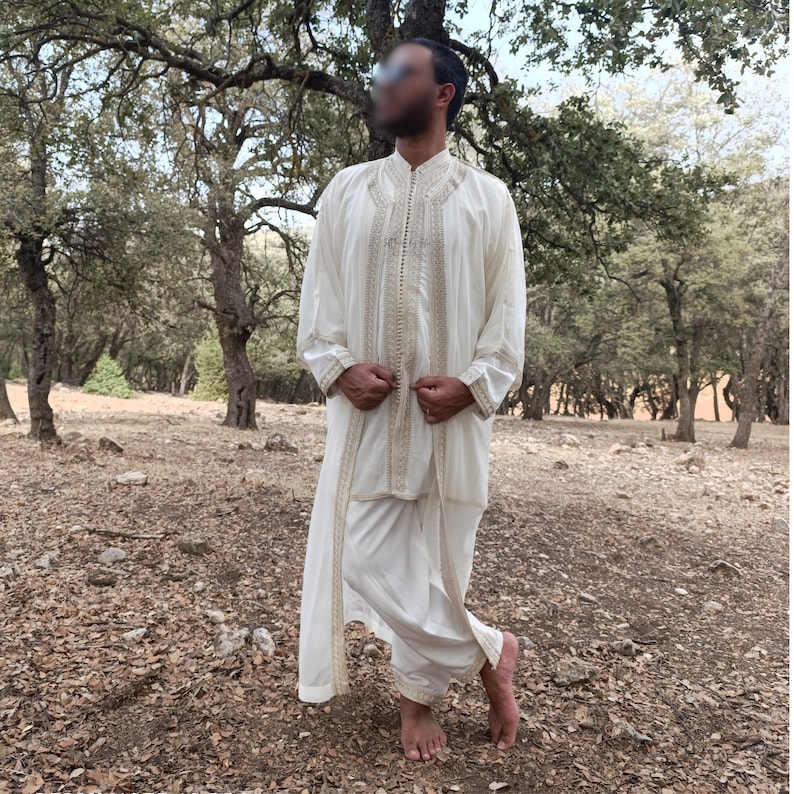 May include: A white long-sleeved robe with gold embroidery and a matching white pant. The robe is open in the front and the person is wearing it with the sleeves rolled up.